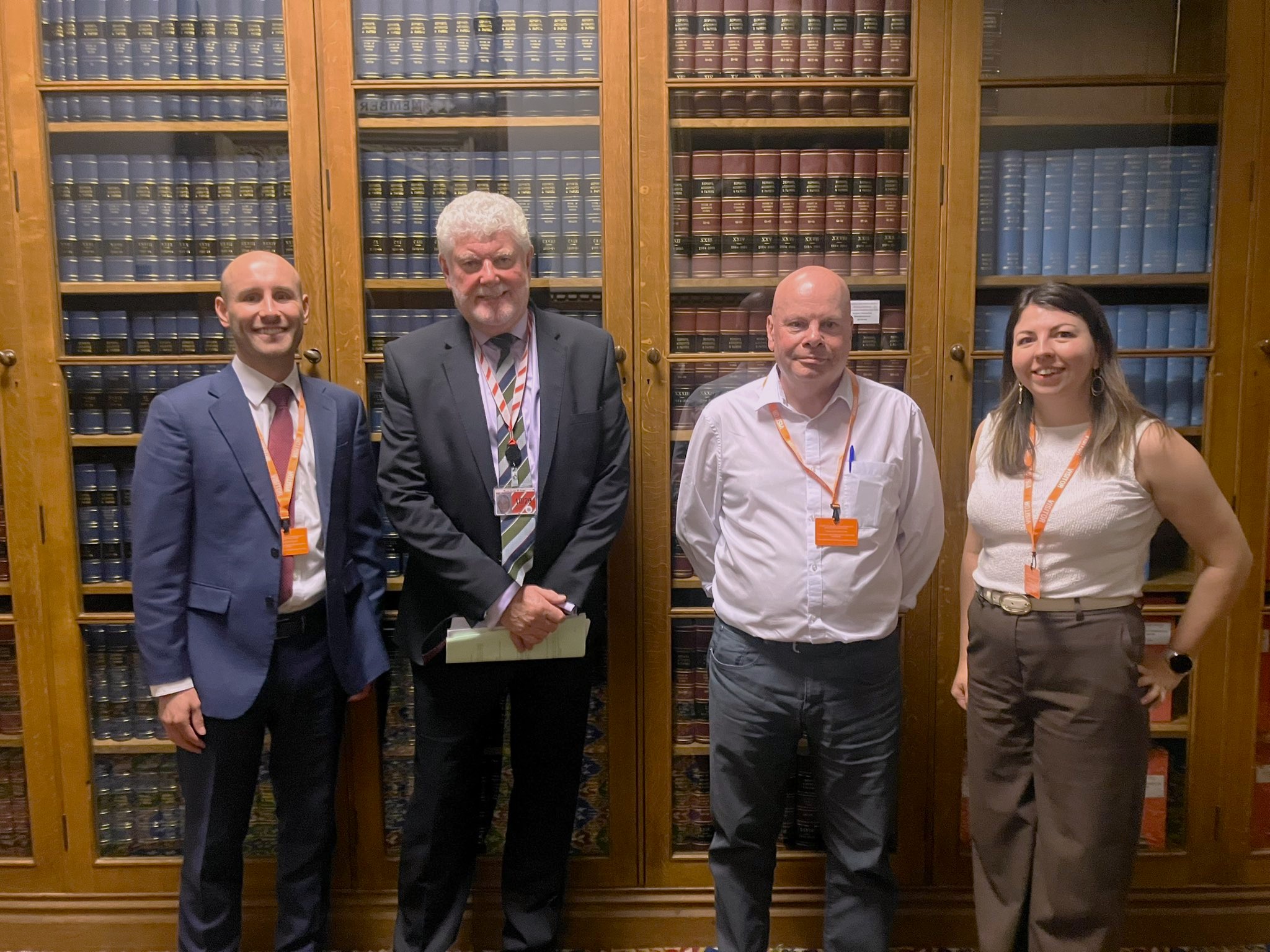 Photo of three men and a woman, smiling at the camera in front of a floor to ceiling bookcase of heavy hardbacks: Tom Robinson, Lord Davies of Gower, Paul Ellsmore, Victoria Parrett